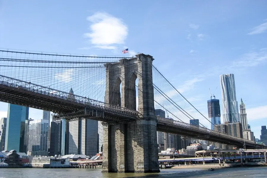 The image shows the Brooklyn Bridge spanning across the East River with the Manhattan skyline in the background on a clear day.