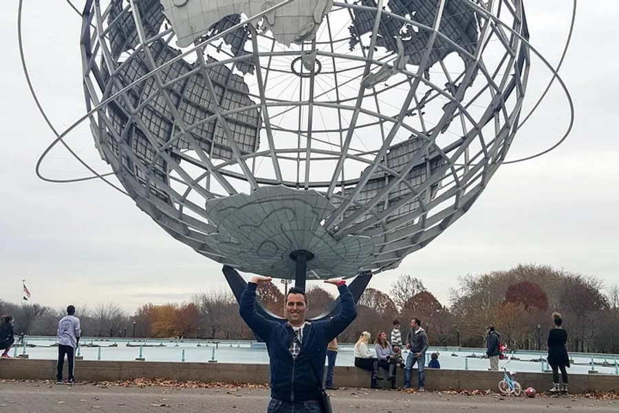 A person is posing with their hands up as if holding the Unisphere, a large globe structure, at Flushing Meadows-Corona Park in New York.