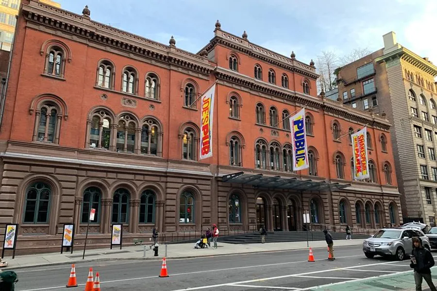 The image shows a large, ornate red brick building with multiple arched windows and a series of colorful banners hanging from its facade, situated on a city street with pedestrians and vehicles nearby.