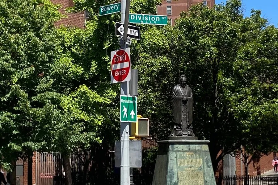 The image features a collection of street signs at an intersection with a statue in the background, surrounded by lush green trees.