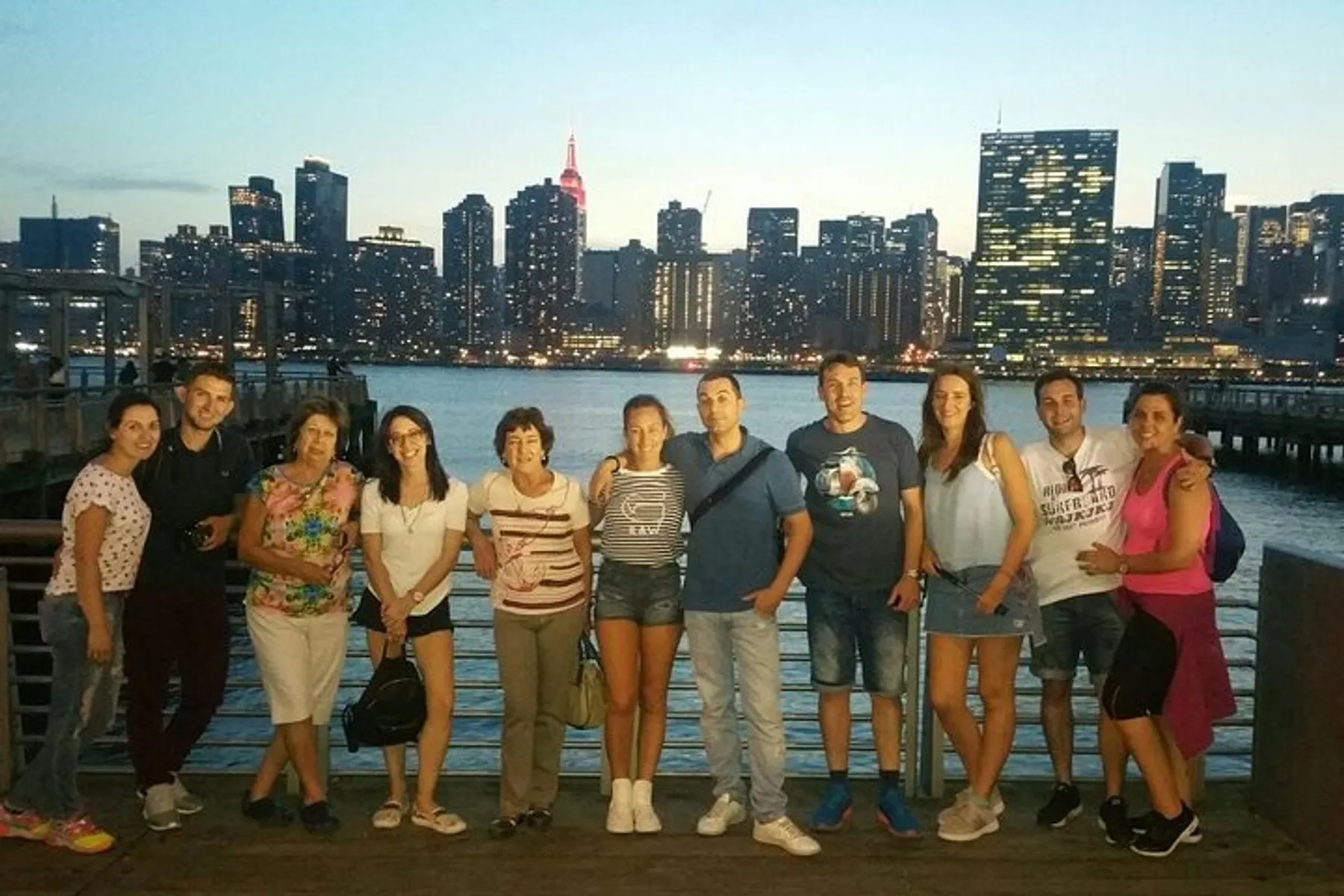 A group of people is posing for a photo on a waterfront with a city skyline illuminated at dusk in the background.