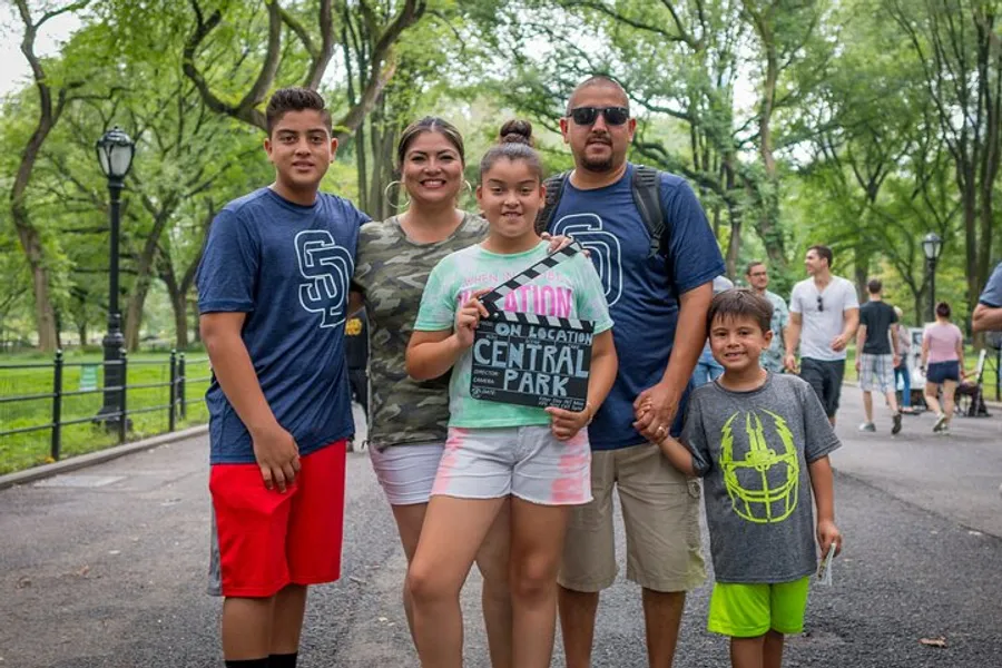 A happy family of five is posing for a photo while enjoying a day out in what appears to be Central Park, with the youngest member holding a guidebook.