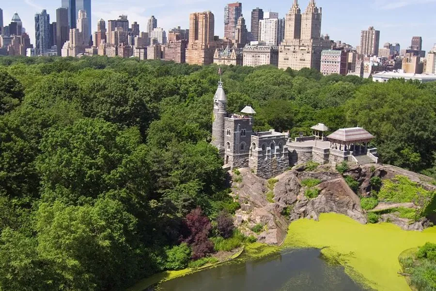 The image captures a historic stone building overlooks a vibrant green pond within a lush park, with the skyline of a bustling city in the background.