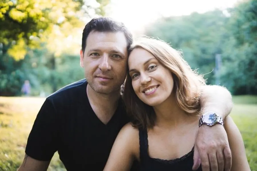 A man and a woman are smiling and posing closely together in a sunlit park.