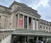 The image shows the exterior of a grand neoclassical building with large banners advertising exhibitions indicative of a museum or cultural institution