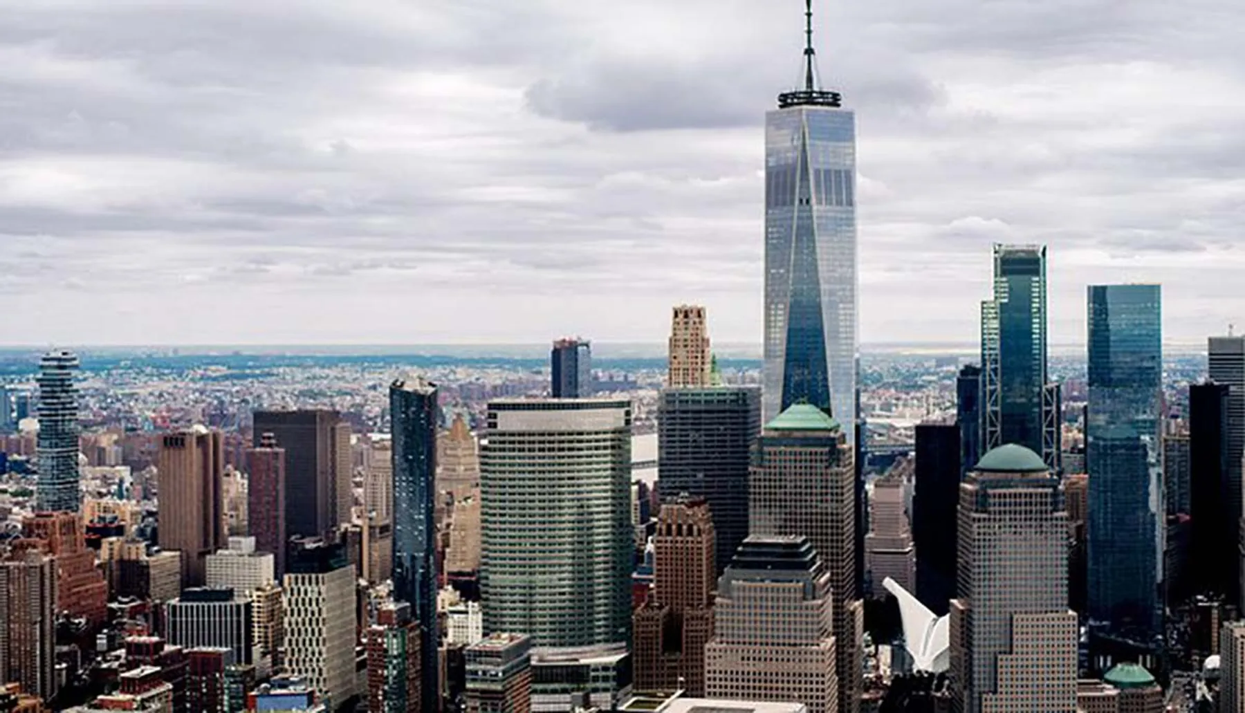 The image is an aerial view of a dense urban skyline dominated by the One World Trade Center amidst numerous skyscrapers, likely depicting Lower Manhattan in New York City.