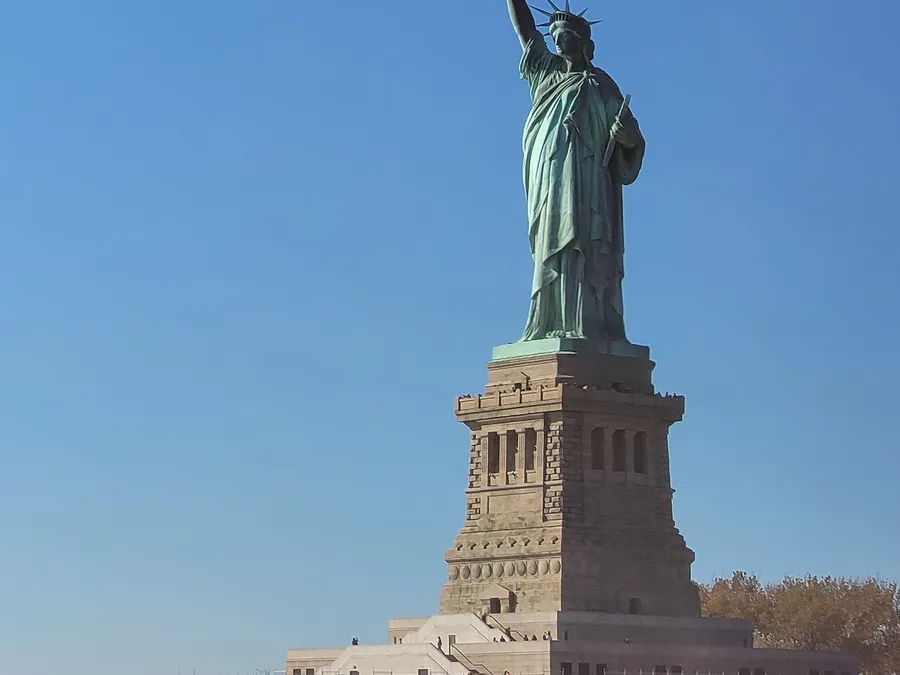 The image shows the Statue of Liberty against a clear blue sky, standing tall on its pedestal.