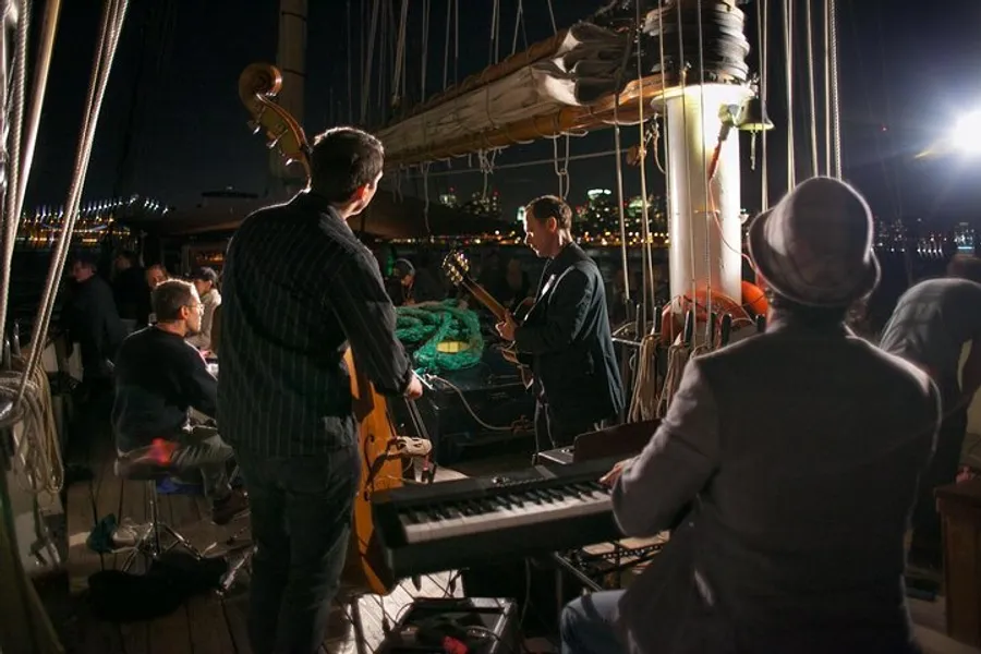 A group of musicians is performing on the deck of a boat at night with city lights in the background.