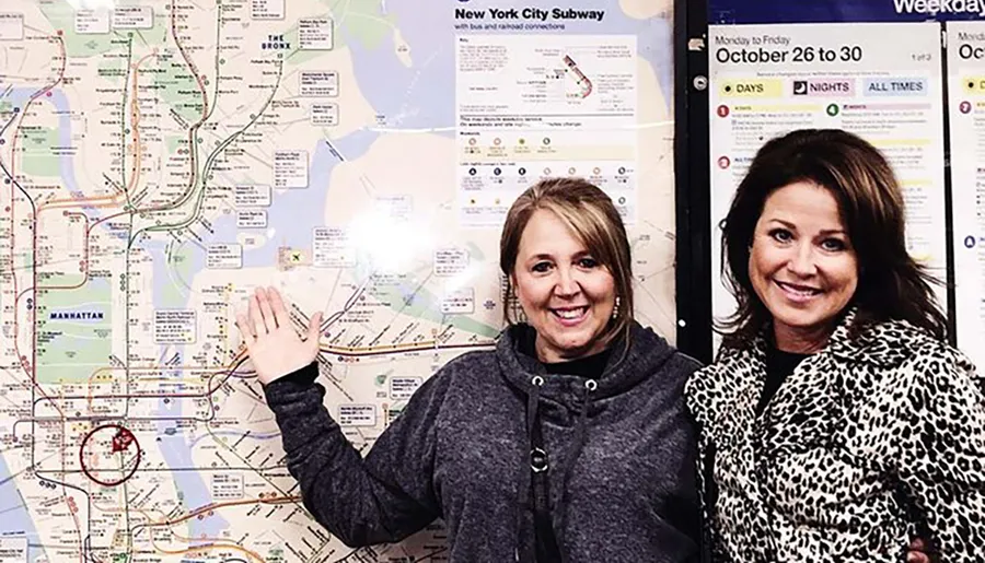 Two smiling women pose in front of a large map of the New York City subway system.