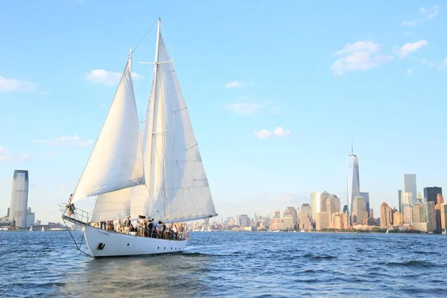 A sailboat filled with people is cruising on calm waters with a backdrop of a modern city skyline.