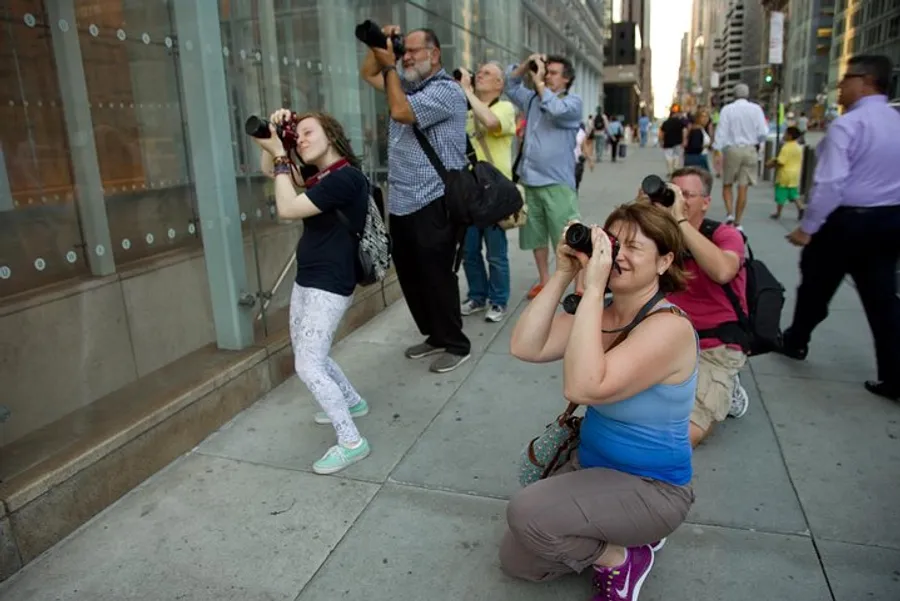 Several individuals are photographing something off-camera on a city sidewalk, some using unique stances to capture their shot.