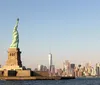 The Statue of Liberty stands prominently in the foreground with the Manhattan skyline including One World Trade Center in the background under a clear sky