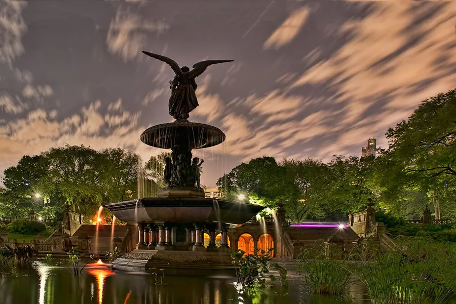 A long-exposure photograph captures the dynamic movement of clouds over the lit-up Bethesda Fountain in Central Park at night.