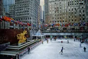 The image showcases an outdoor ice-skating rink surrounded by tall buildings, with a golden statue to the left, a row of international flags, and people skating or walking around the rink.