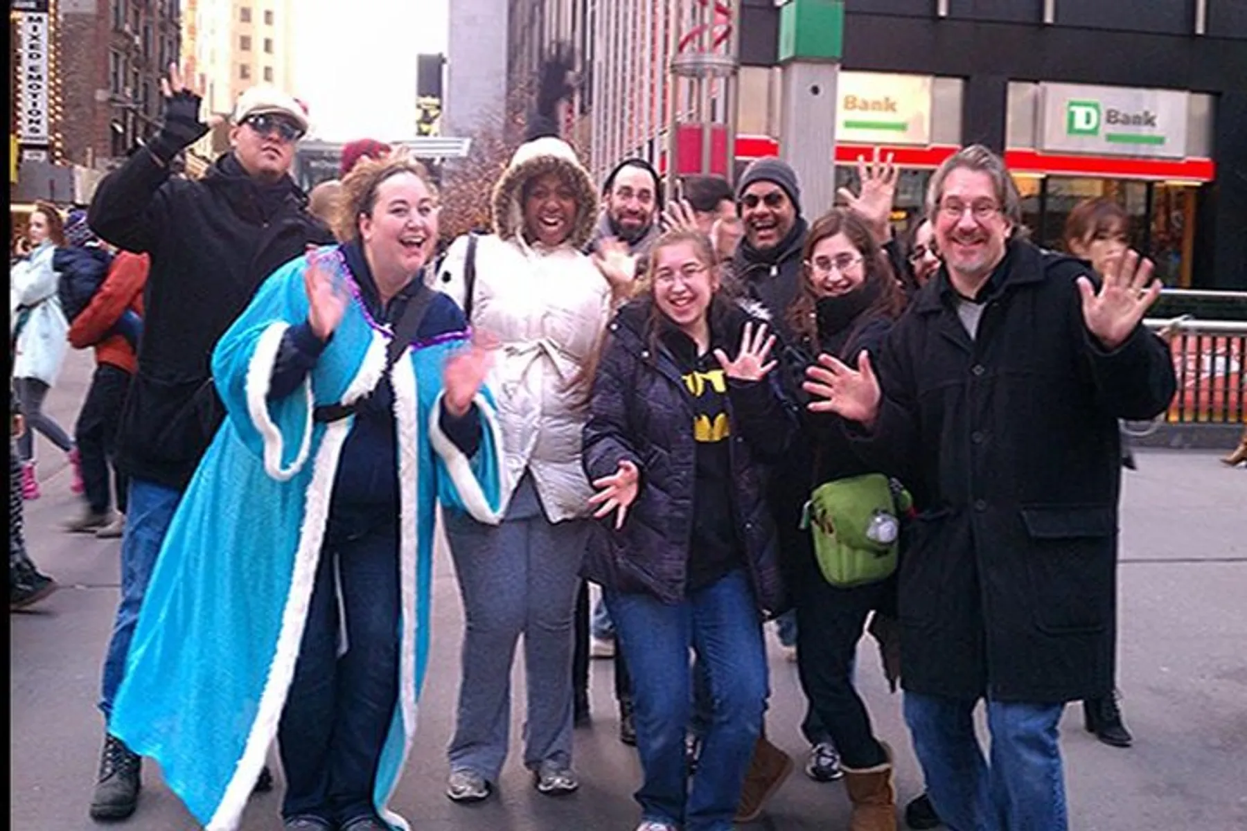 A group of cheerful people posing for a photo on a busy city sidewalk, with some raising their hands or making gestures to the camera.