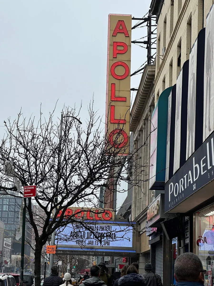 This image shows people walking by the iconic Apollo Theater marquee with neon signage in an urban setting.