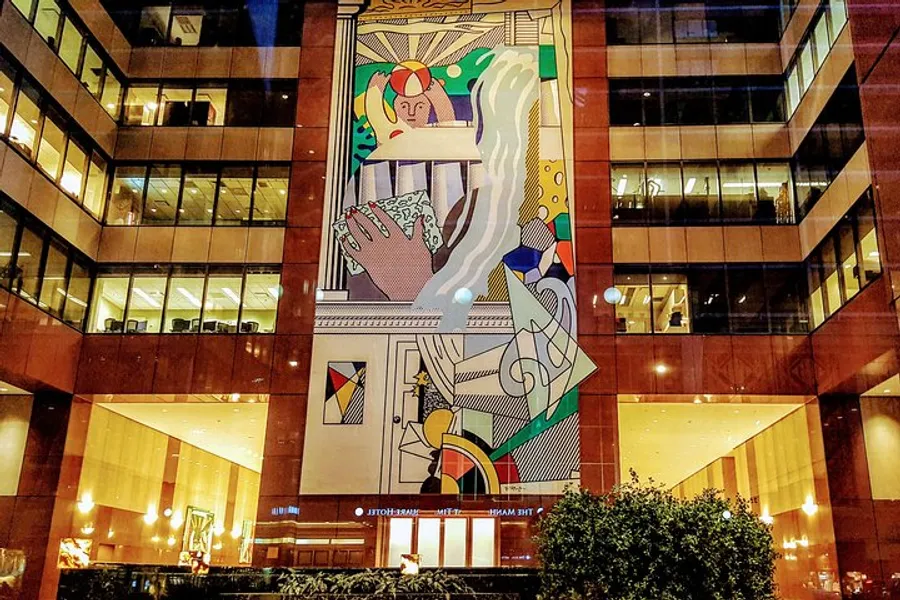 The image shows a colorful mural with abstract and figurative elements displayed on a building's facade at night, accentuated by indoor lighting.