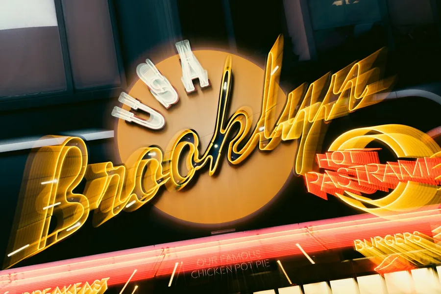 The image shows a neon sign that reads Brooklyn with a motion blur effect, and features words like USA, Hot Pastrami, Burgers, and Our Famous Chicken Pot Pie.