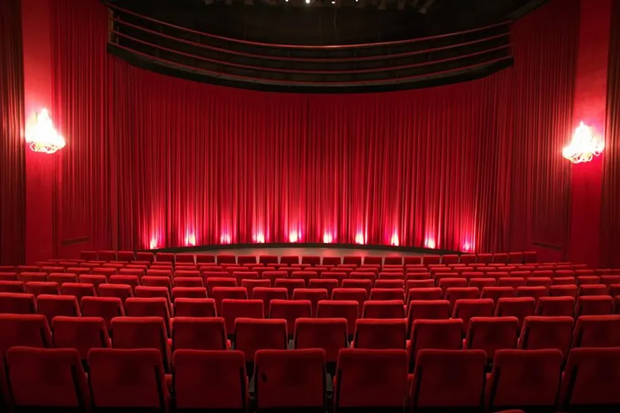 An empty theater with red seats facing a closed red curtain on the stage and ambient lighting.