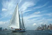 A sailboat is cruising on the water with a city skyline in the background under a partly cloudy sky.