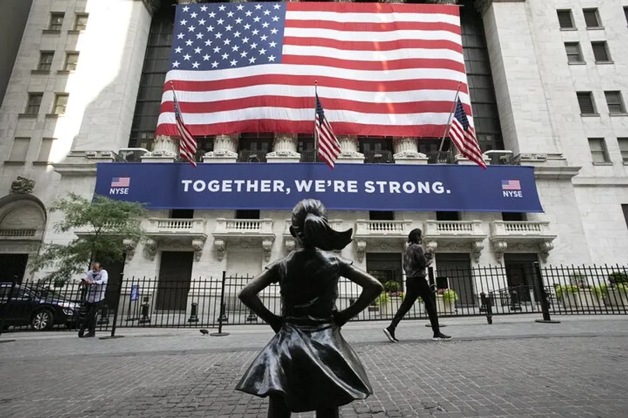 A statue of a girl stands defiantly in front of the New York Stock Exchange, which is adorned with an American flag and a banner saying TOGETHER, WE'RE STRONG.