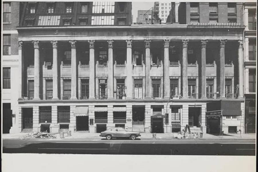 The image shows a vintage black-and-white photograph of an old building with classical columns and evident signs of wear, located in an urban environment with a single classic car parked in front.