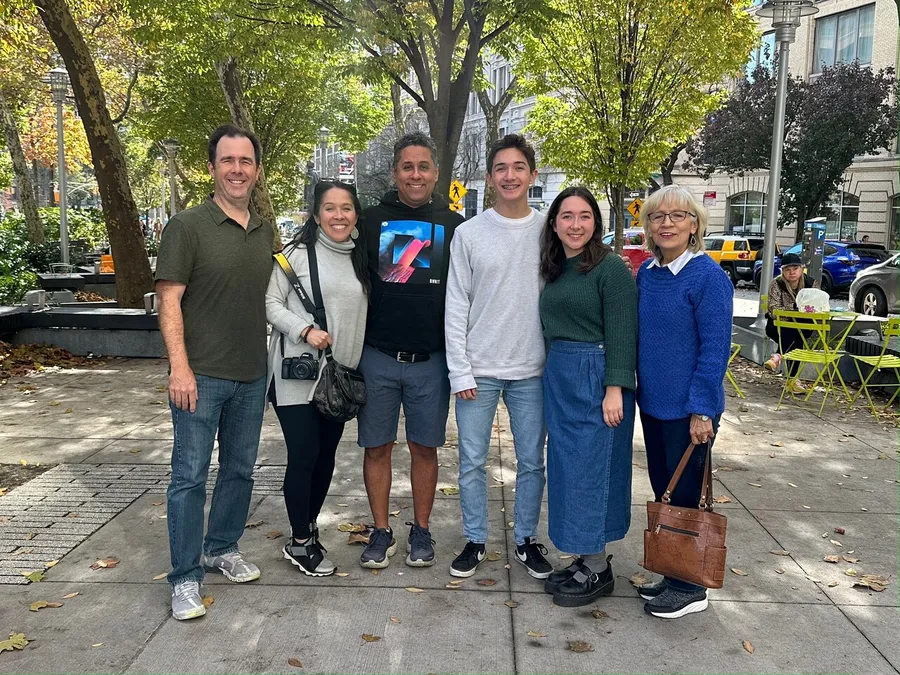 A group of six people are smiling for a photo on a sunny day with trees and a city street in the background.