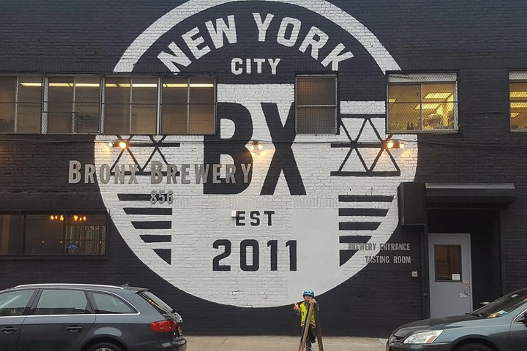 The image shows the exterior of a building painted black with a large white circular logo that reads New York City BX Bronx Brewery EST 2011, indicating the establishment is a brewery in the Bronx, New York City, established in 2011, with a person wearing a helmet standing in front of it.