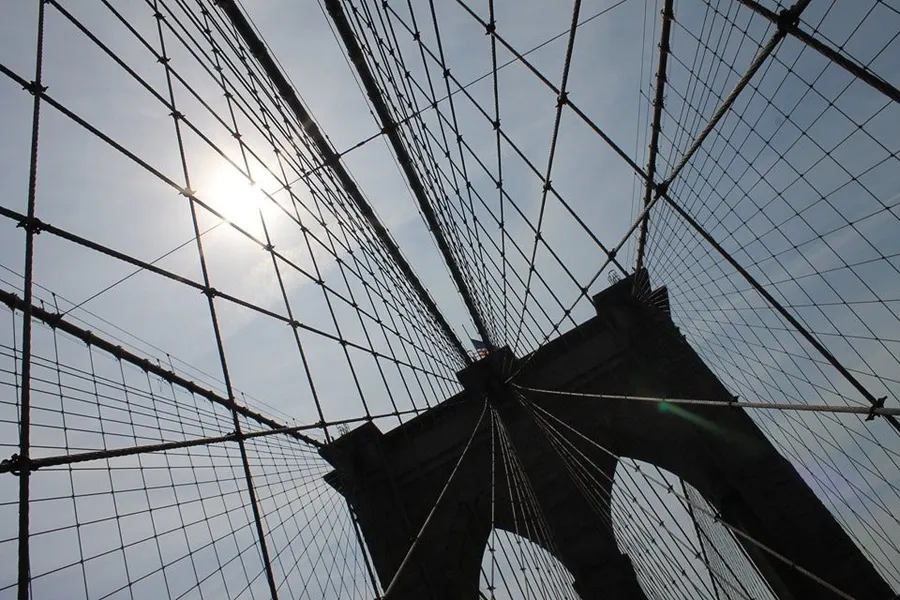 The image captures a striking view of the sun peeking through the network of cables of a suspension bridge against a clear sky.