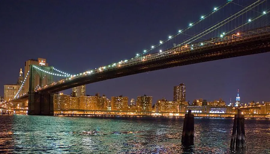The image shows the illuminated Brooklyn Bridge at night with the East River in the foreground and the city skyline in the background.