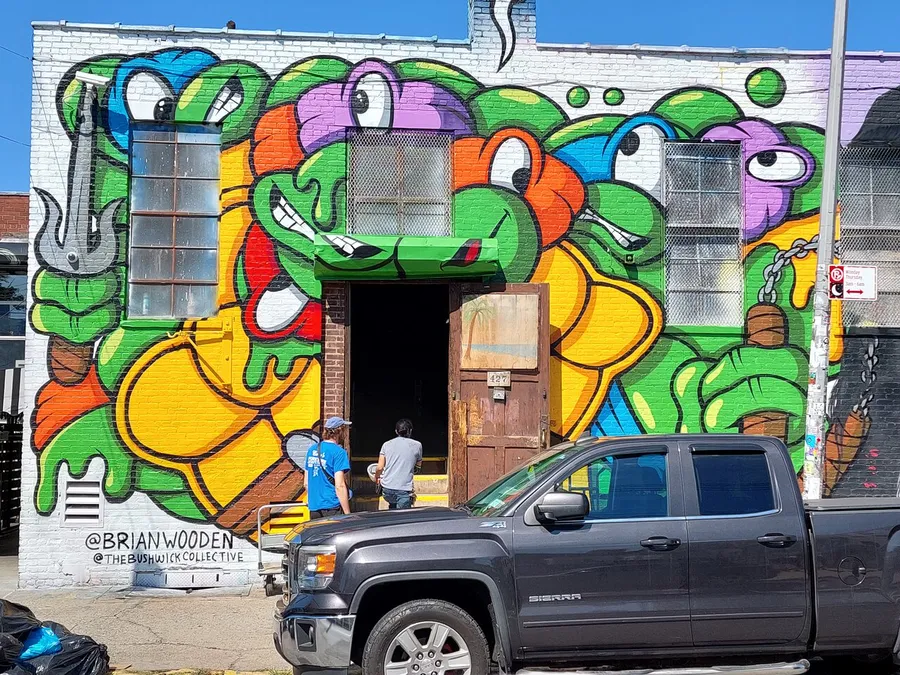 The image shows a vibrant street mural featuring colorful, cartoonish snake characters on a building's exterior, with two people and a truck visible in front of the artwork.