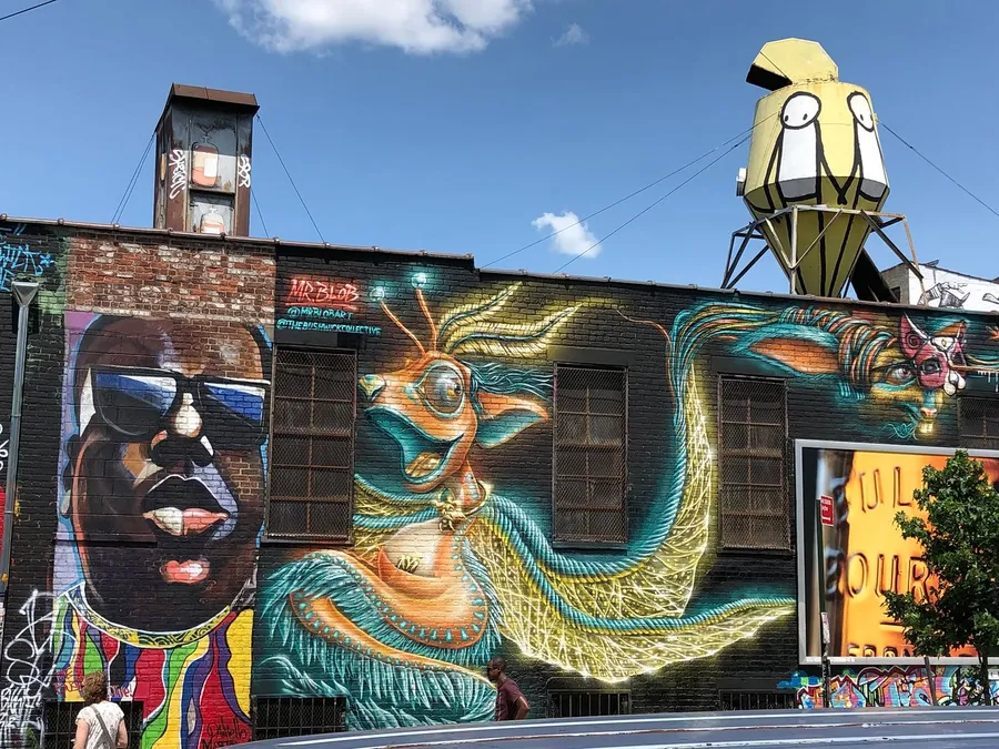 The image shows a vibrant street art mural featuring various colorful and stylized figures, with a whimsical sculpture on a water tower above the building.