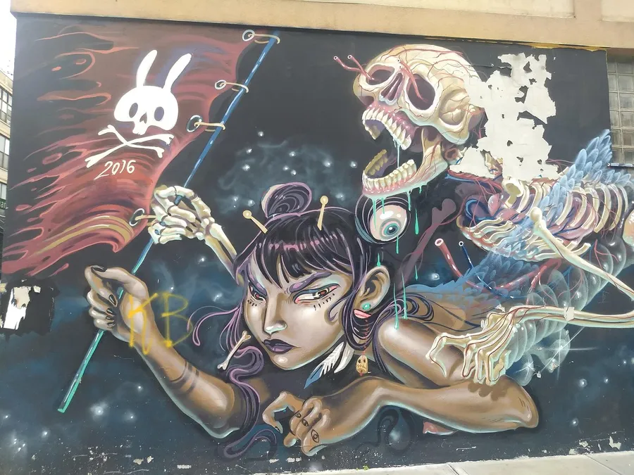 This is a vibrant street art mural depicting a woman with puppet strings controlling a skeleton and a skull marked with a bunny symbol, blending elements of fantasy and surrealism.