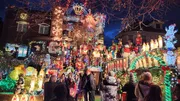 A group of people gathers in front of an elaborately decorated house with an abundance of colorful Christmas lights and festive ornaments, creating a vibrant holiday atmosphere.