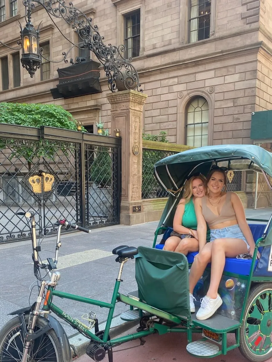 Two people are smiling while seated in a green pedicab parked in front of an elegant building with ironwork detailing.
