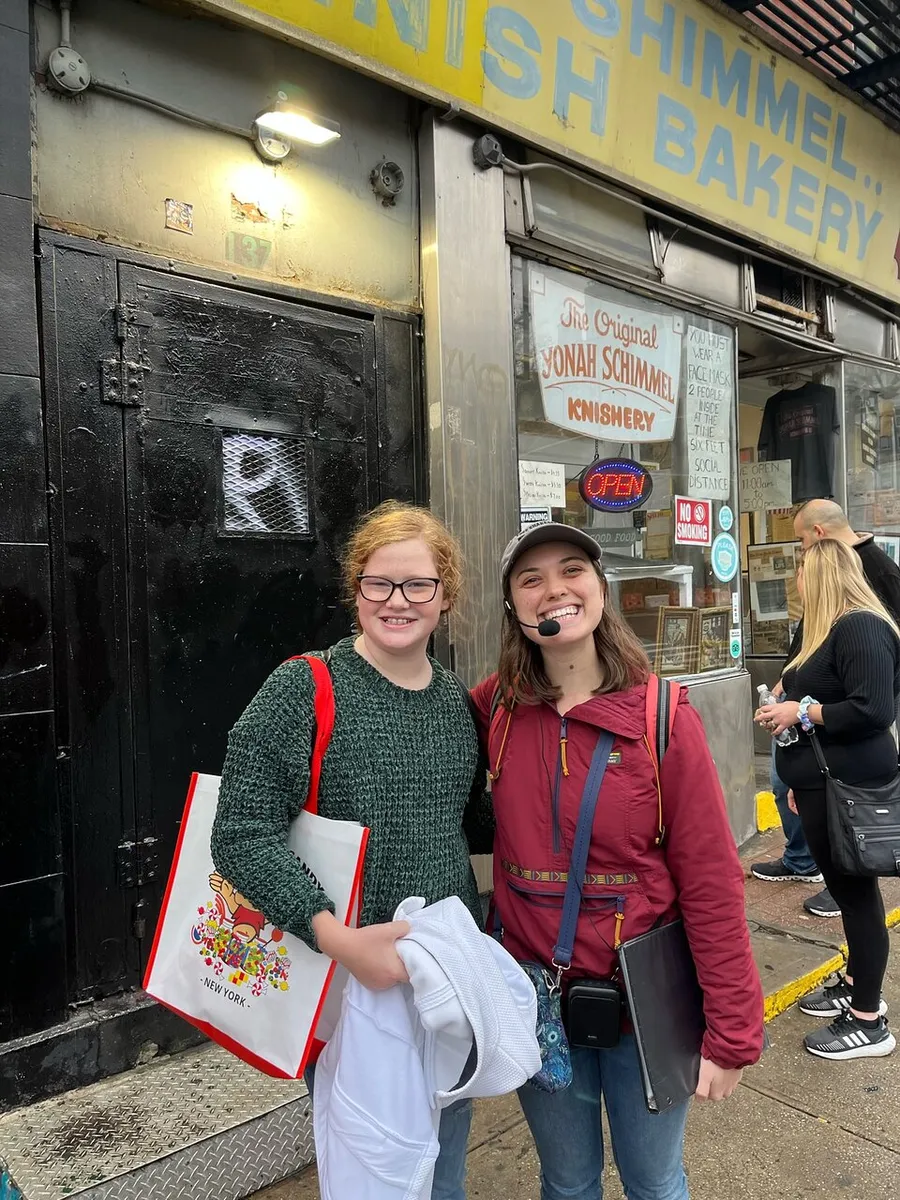 Two smiling young individuals are standing in front of the Yonah Schimmel Knish Bakery, possibly after having visited the shop.