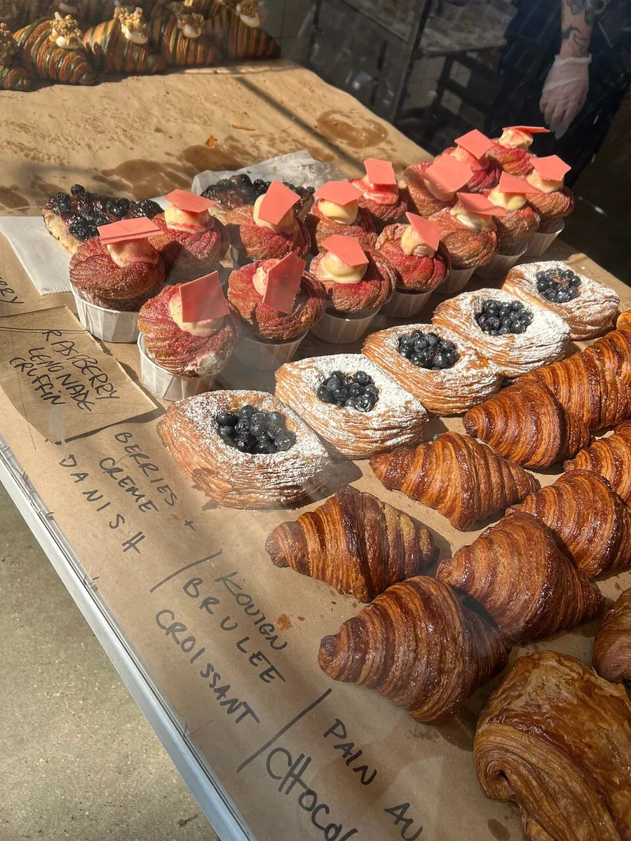 The image shows a variety of freshly baked pastries displayed in a bakery, with items labeled such as blackberry crème fraiche craham and kouign brulee croissant.