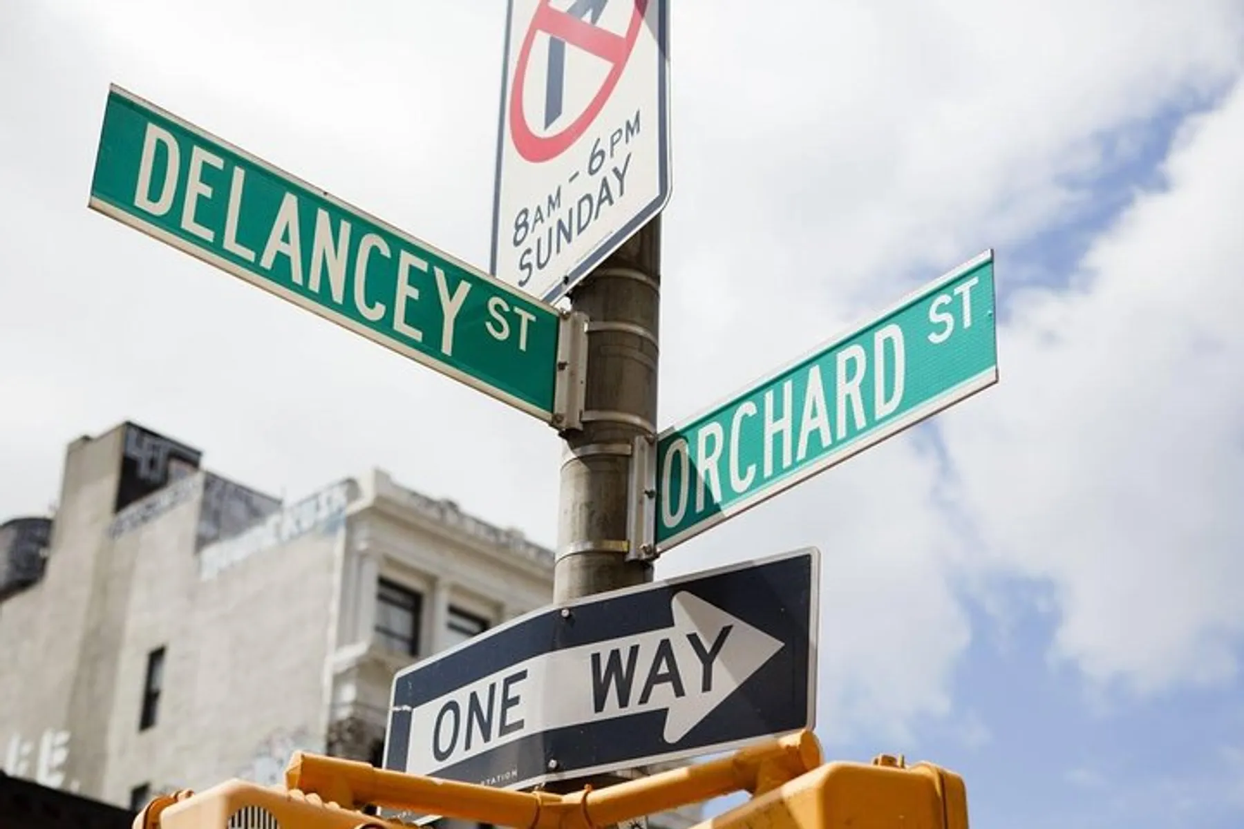 The image shows street signs at the intersection of Delancey Street and Orchard Street with a 'No Parking' sign and a 'One Way' sign attached to a traffic light pole against a partly cloudy sky.