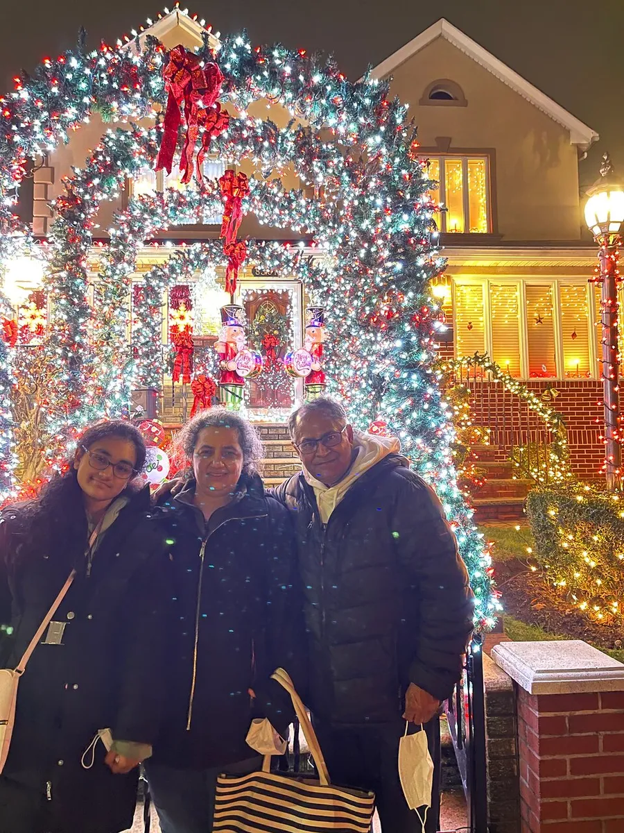 Three people are posing for a photo in front of a festively decorated house with a large, illuminated Christmas wreath archway.