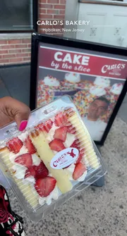 A person is holding a slice of strawberry-topped cake in a clear container in front of a Carlo's Bakery sign in Hoboken, New Jersey.