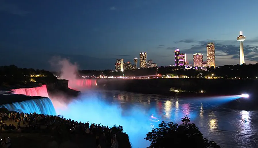 The image captures a night view of Niagara Falls with illuminated water in blue and red, overlooking a lit city skyline and gathering of spectators.