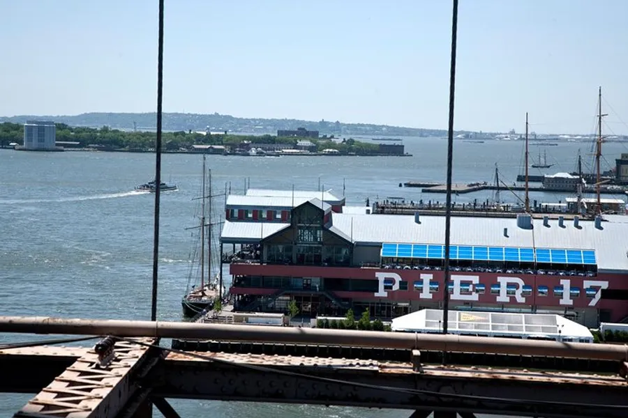 The image shows a waterfront view of Pier 17 with a ship docked nearby, taken from above and framed by cables, likely from a bridge.