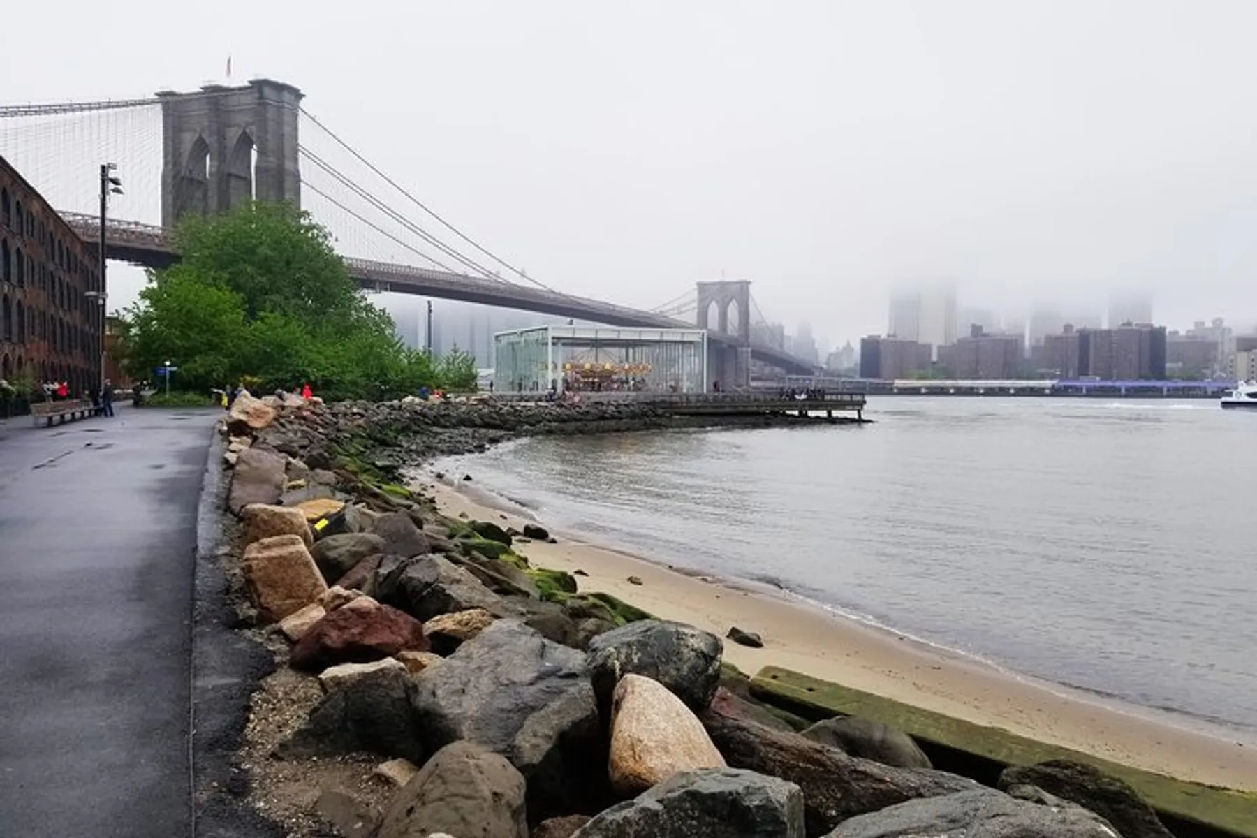 The image features the Brooklyn Bridge on a foggy day, with a view from the riverbank showcasing a carousel structure and the mist-obscured skyline of Manhattan in the background.