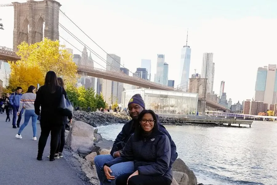 Two people are smiling for the camera while seated on rocks near a waterfront park with the Brooklyn Bridge and the Manhattan skyline in the background.