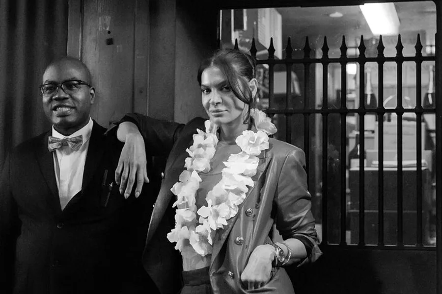 A man in a tuxedo with a bow tie and glasses stands next to a woman wearing a lei and a blazer, both of whom are posing with confidence in a monochrome photo.