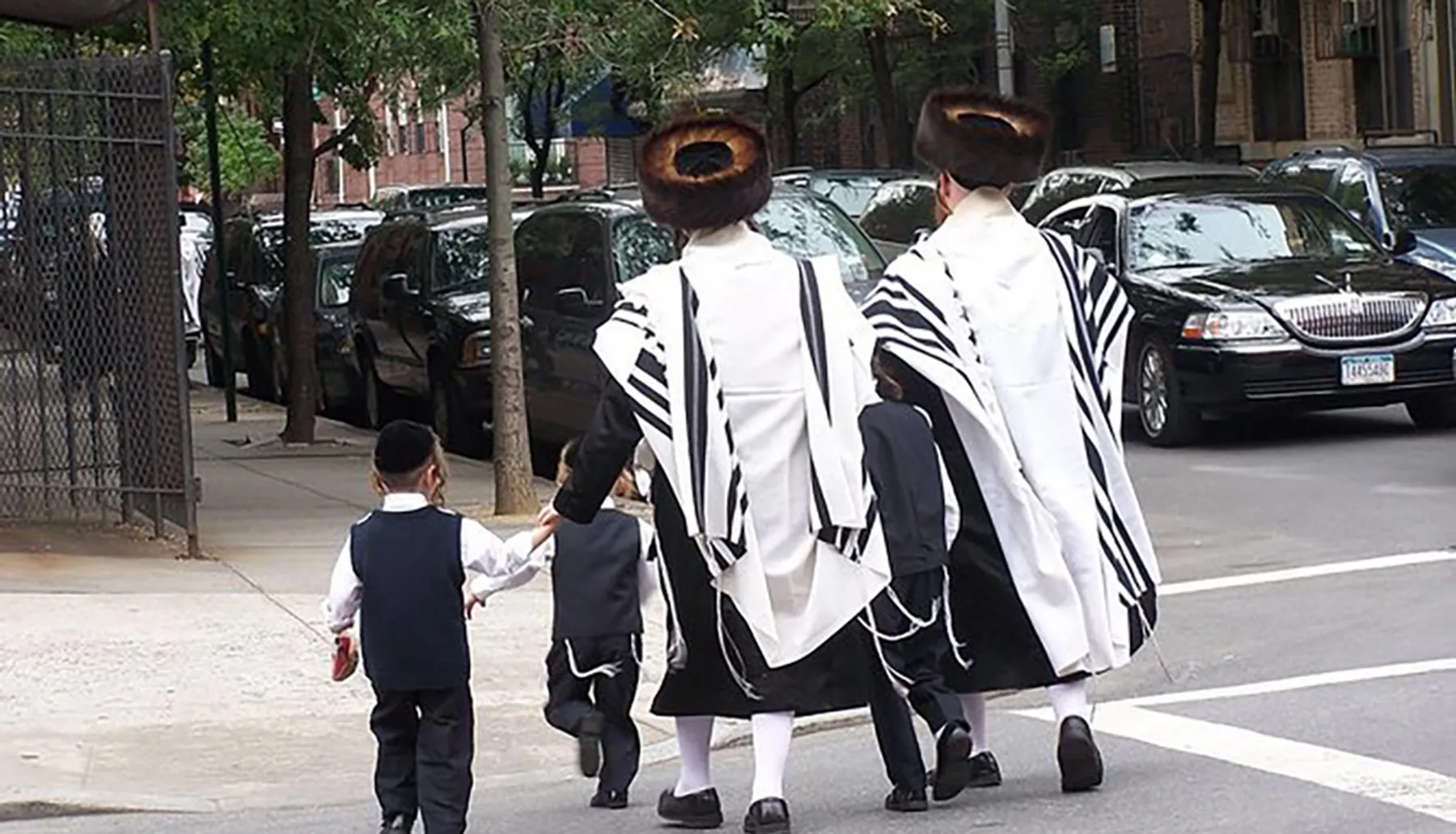 Two adults and two children, dressed in traditional Hasidic Jewish attire with prayer shawls and fur hats, are walking down a city sidewalk.