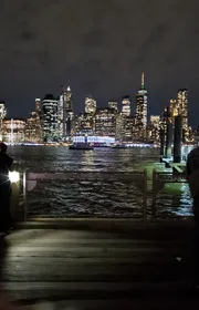 A nighttime view of a brightly lit city skyline seen from a pier with dark waters in the foreground.