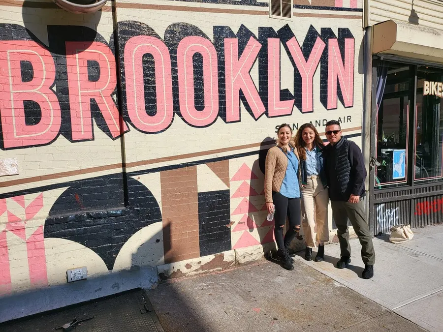 Three people are smiling and posing in front of a large mural that spells out BROOKLYN on a sunny day.