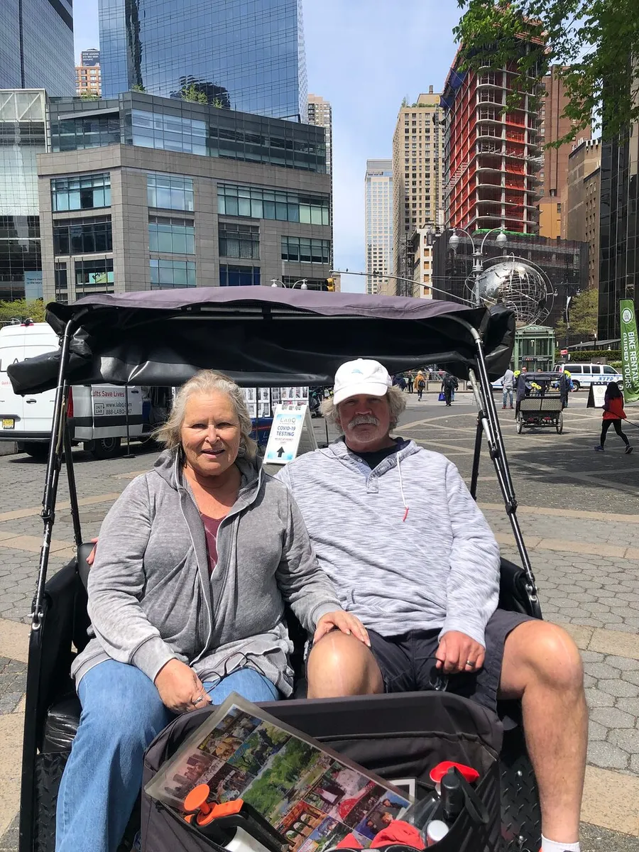 A smiling couple is sitting in a pedicab in an urban setting featuring modern buildings and a globe sculpture in the background.