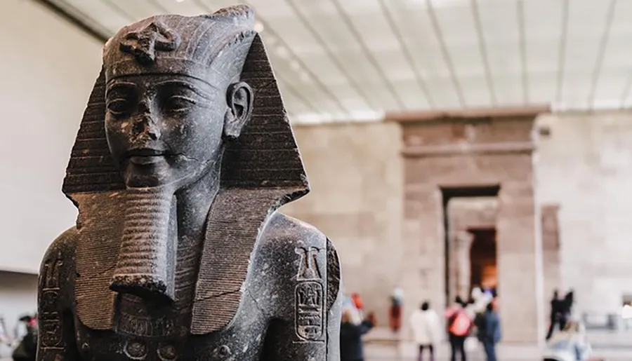 The image features an ancient Egyptian statue of a pharaoh exhibited in a museum with visitors in the background.